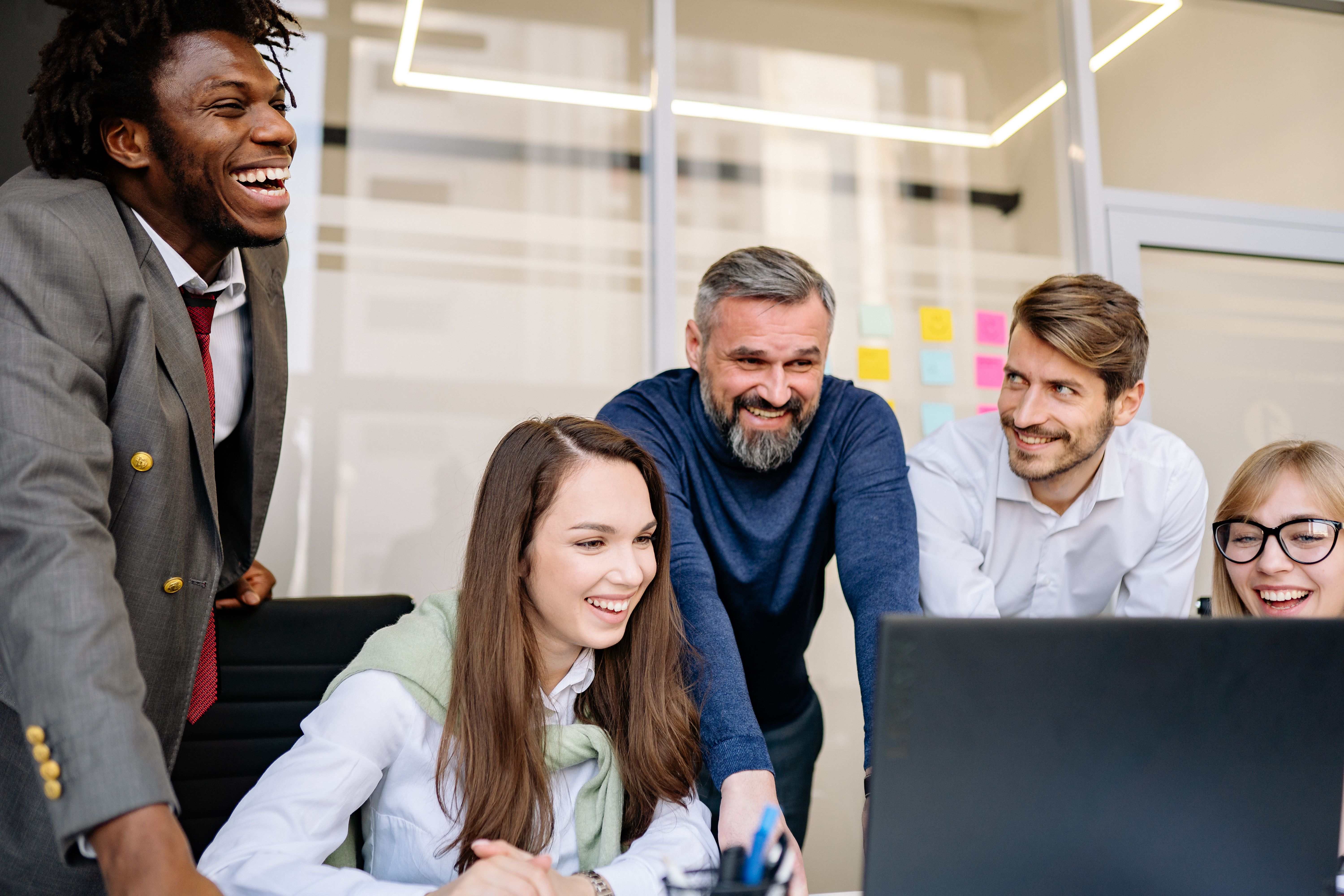 A diverse group of professionals of different races, genders, and ages laughing and collaborating together around a computer in a modern office